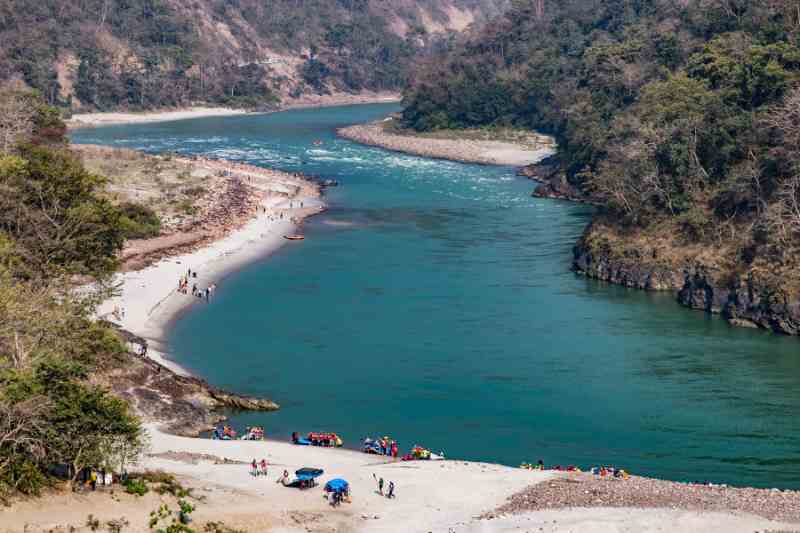 Rio de águas tranquilas e límpidas, margeado por vegetação e colinas verdes. Na margem do rio, pessoas desfrutam de atividades ao ar livre.
