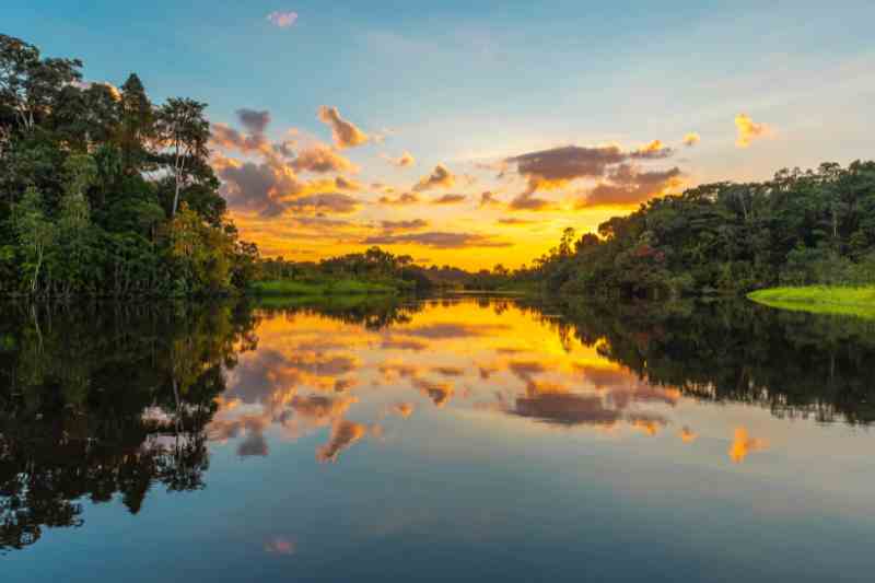 Rio de águas tranquilas, margeado por vegetação natural, reflete o por do sol.