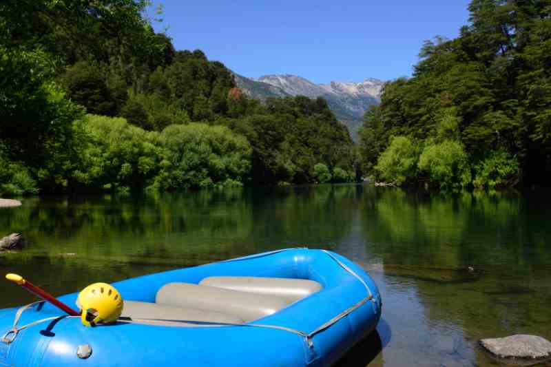 Bote azul, parado às margens de um rio de águas verdes claras, envolto de vegetação densa.