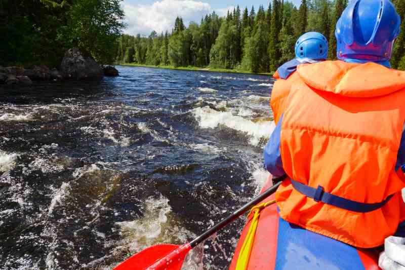 Pessoas em bote vermelho descem rio cercado por pinheiros.