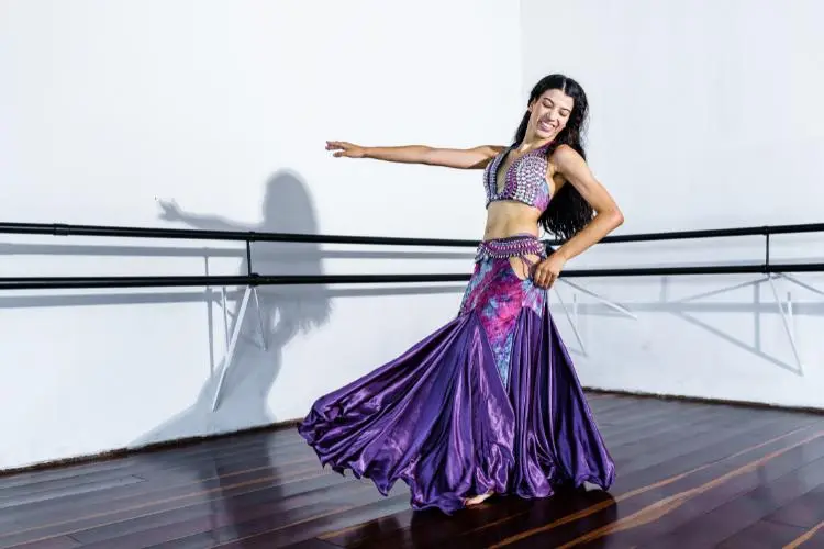 Mulher sorrindo dançando dança do ventre com traje roxo. História, técnicas e significados da dança do ventre.